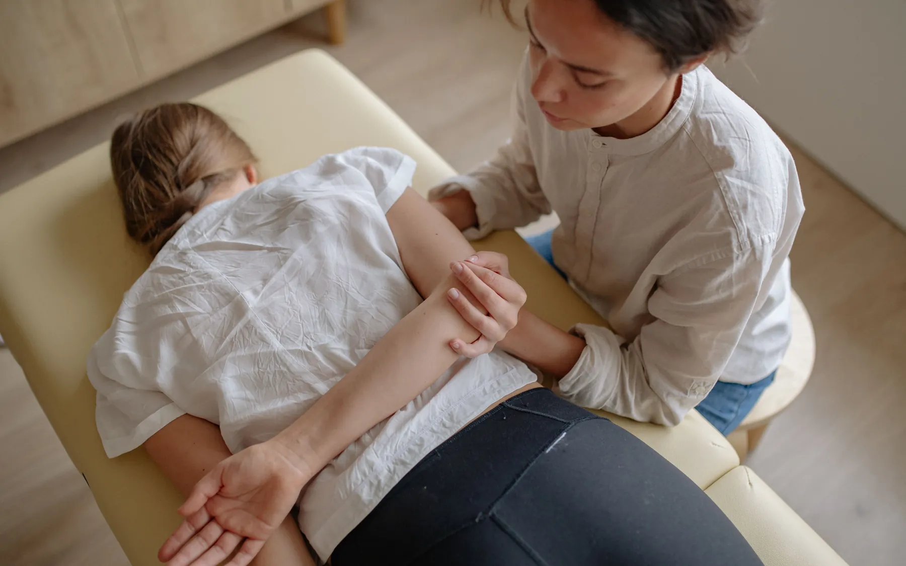 A chiropractor works on the back and arm of a patient lying face down on a treatment table in a sunlit room.