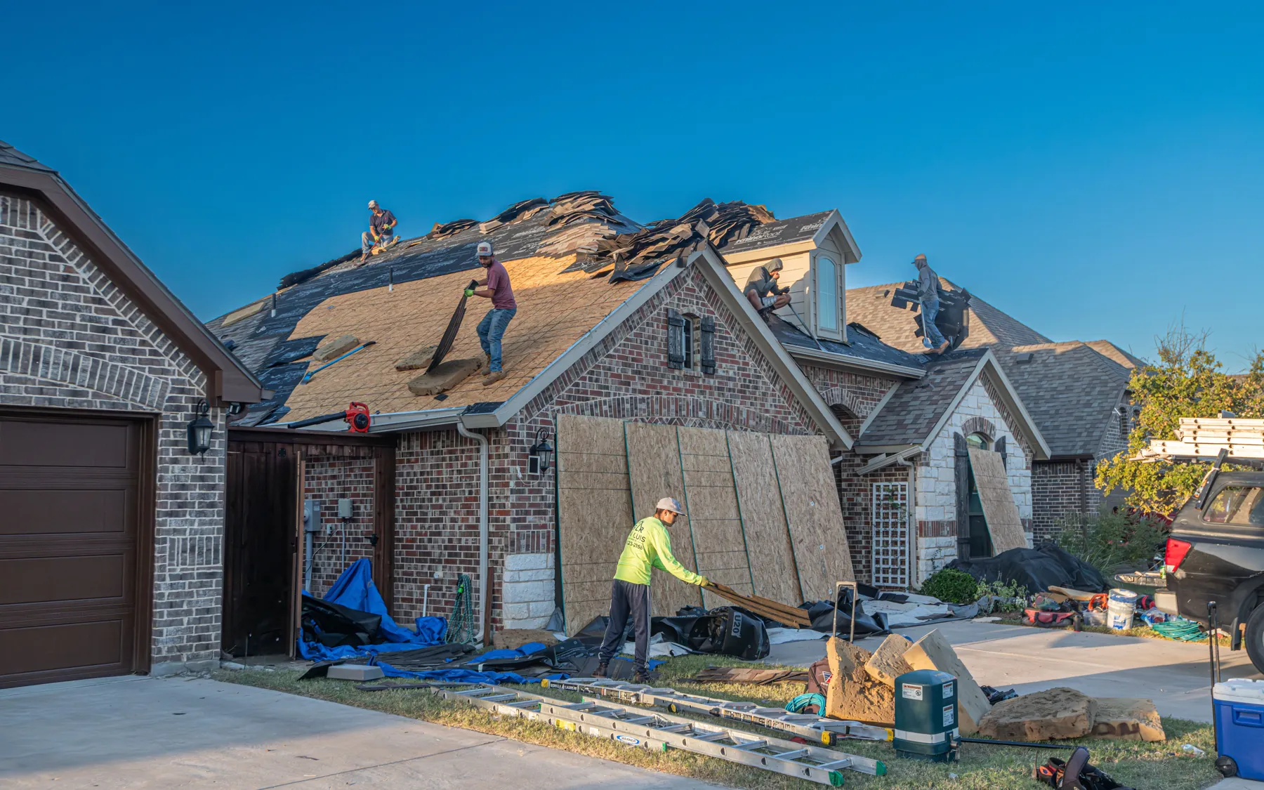 A roofing crew tears off and re-decks the roof of a brick house while a worker carries a sheet of plywood across the lawn.
